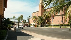 A street view with a large brick building featuring a tower and a church-like structure. Palm trees line the sidewalk, and there are cars parked along the street.