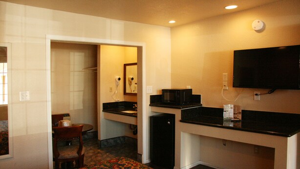 A compact hotel room with a kitchenette, a bed with a floral bedspread, a dining area with a chair, and a television mounted on the wall.