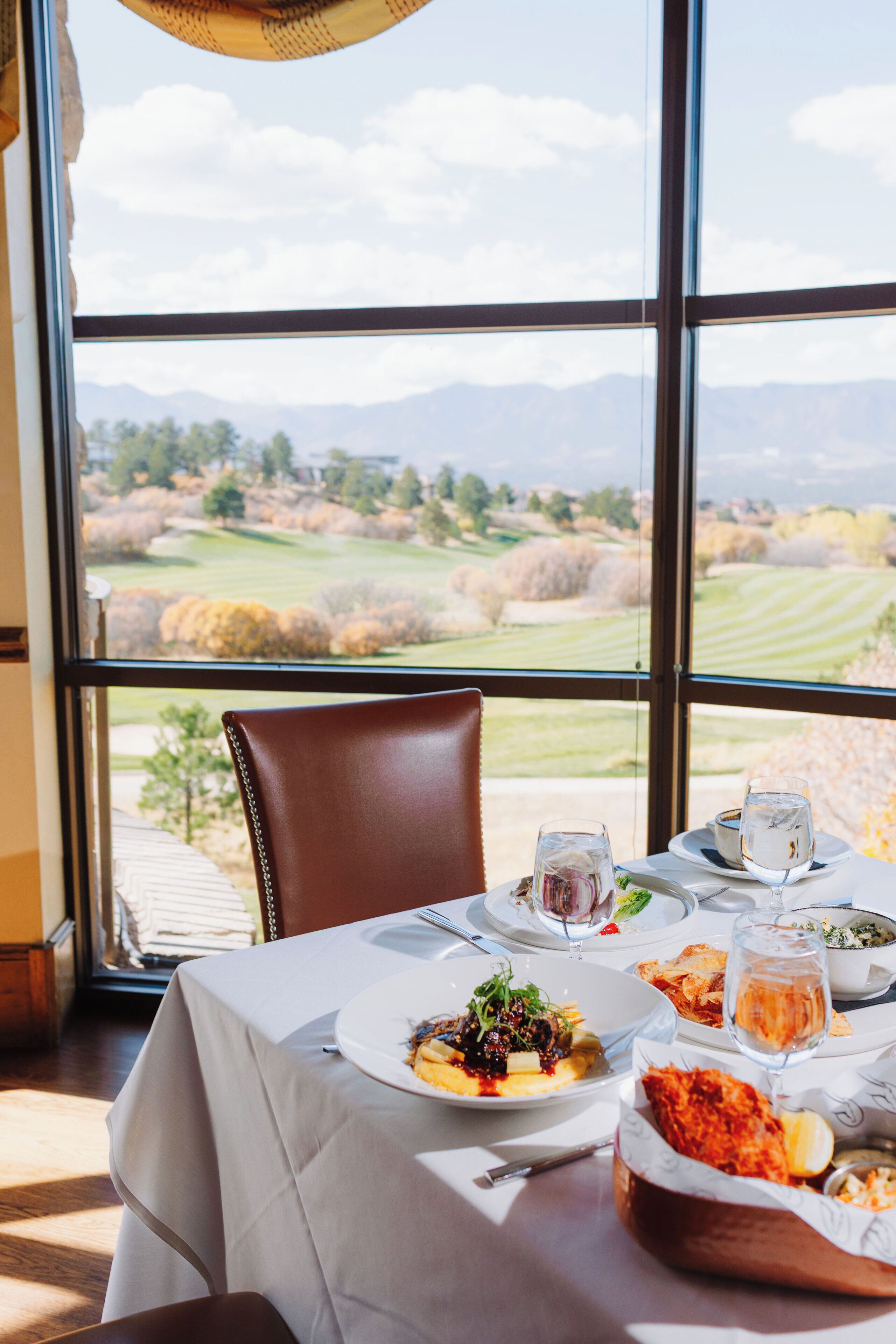 A dining table with plates of food, glasses, and a candle, set against a window with a view of a landscape.