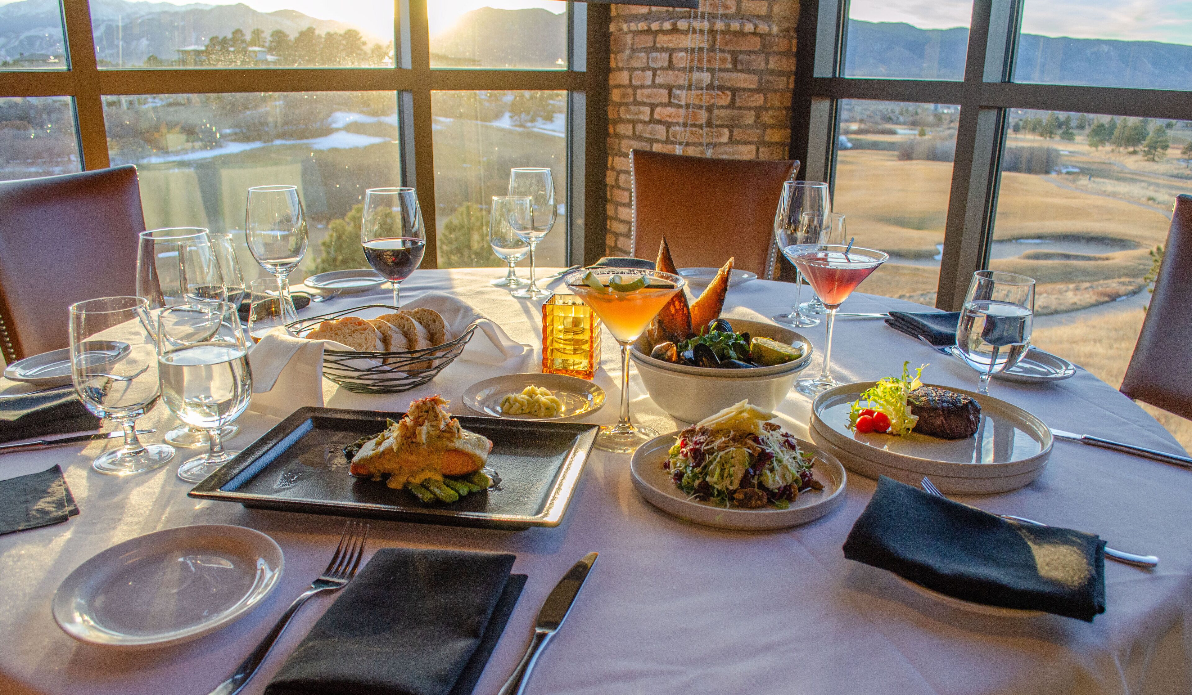 A dining table set with various dishes, glasses, and cutlery, with a view of a landscape through large windows.