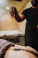 Una persona aplicando un polvo negro en la espalda de otra, en una habitación con pared de piedra y una bañera al fondo.