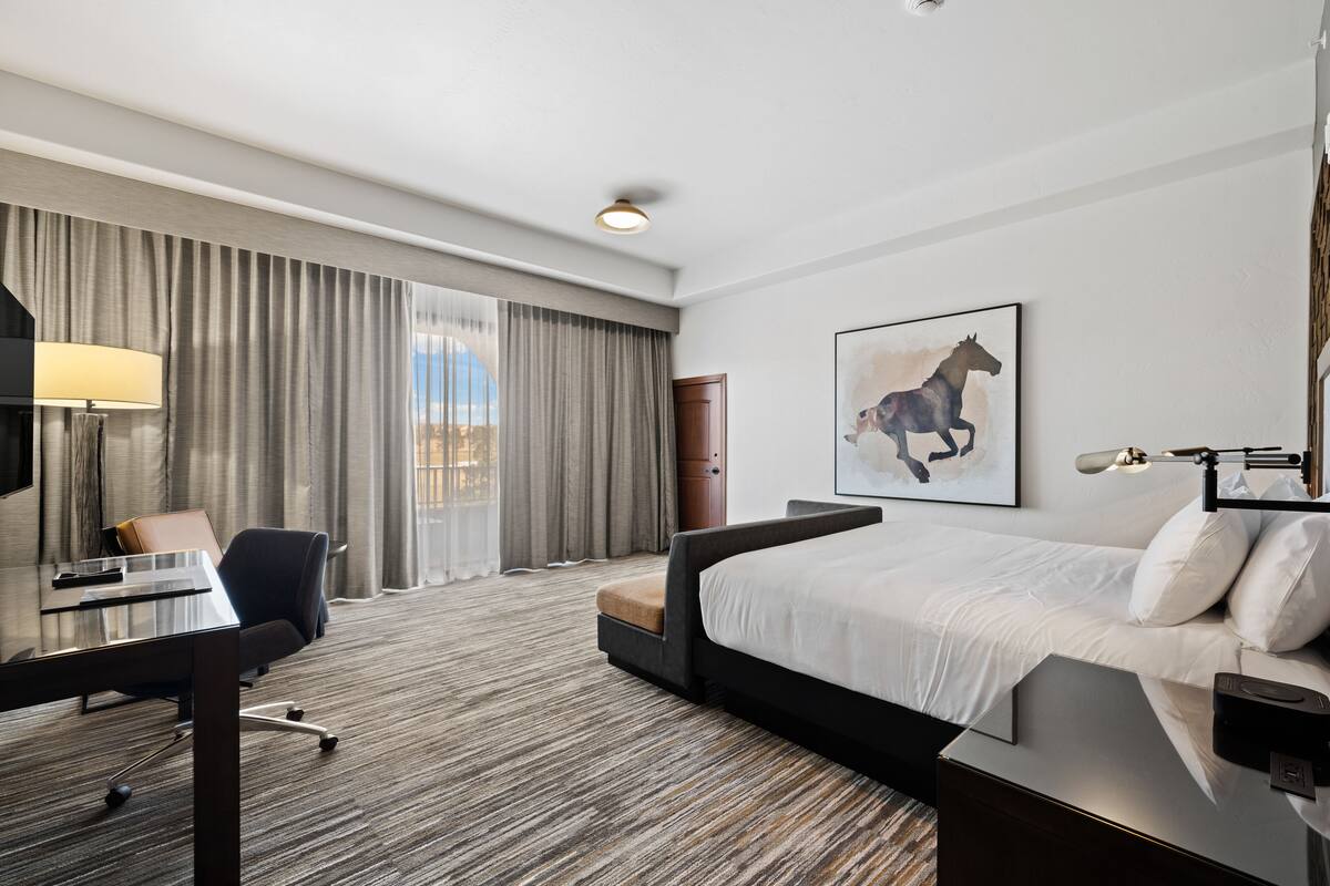 Een hotelkamer met een groot bed, een bureau met stoel, een lamp en een schilderij van een paard aan de muur.