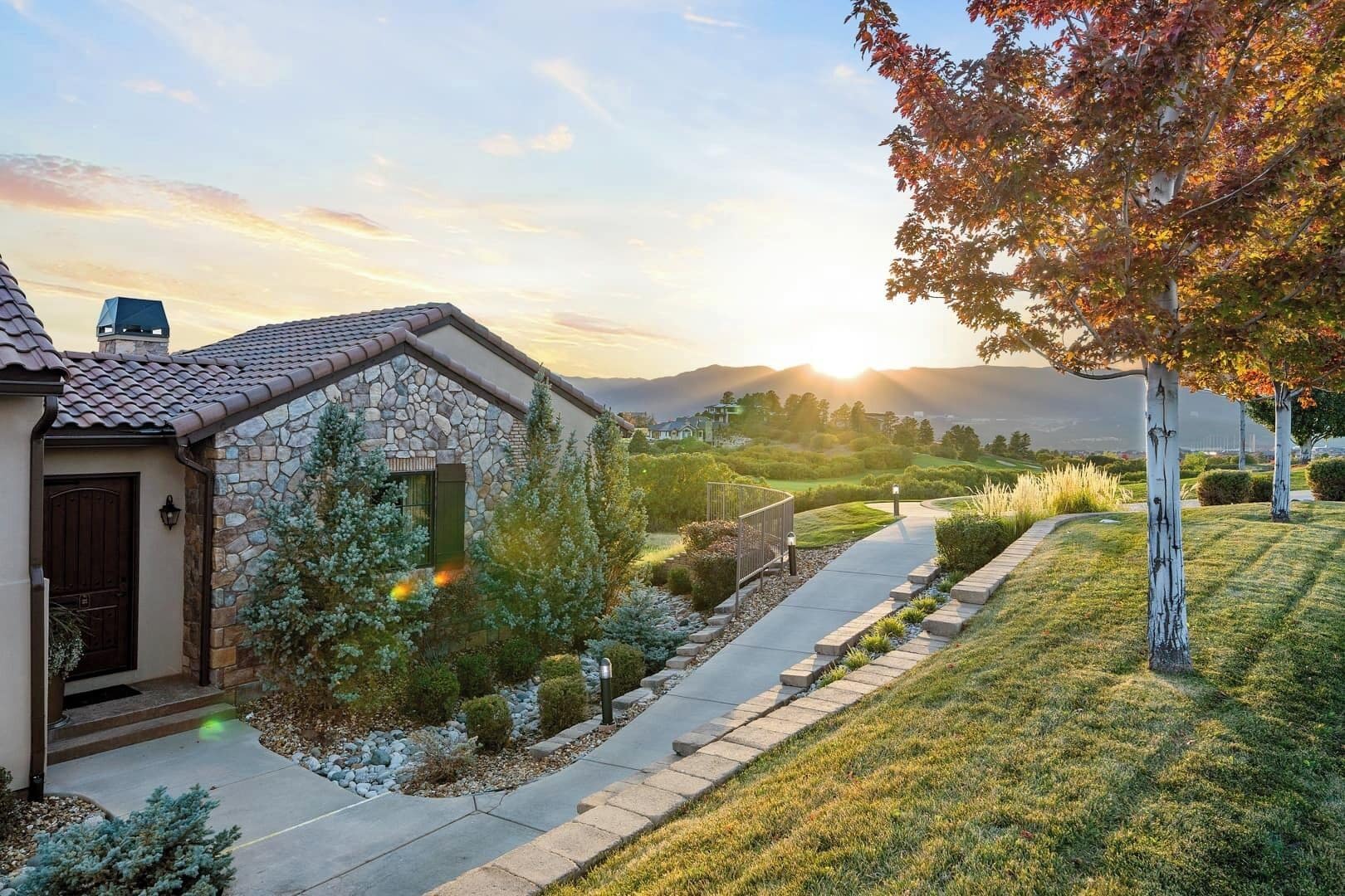 A stone house with a tiled roof, a paved driveway, and a well-maintained lawn with a tree displaying autumn leaves.