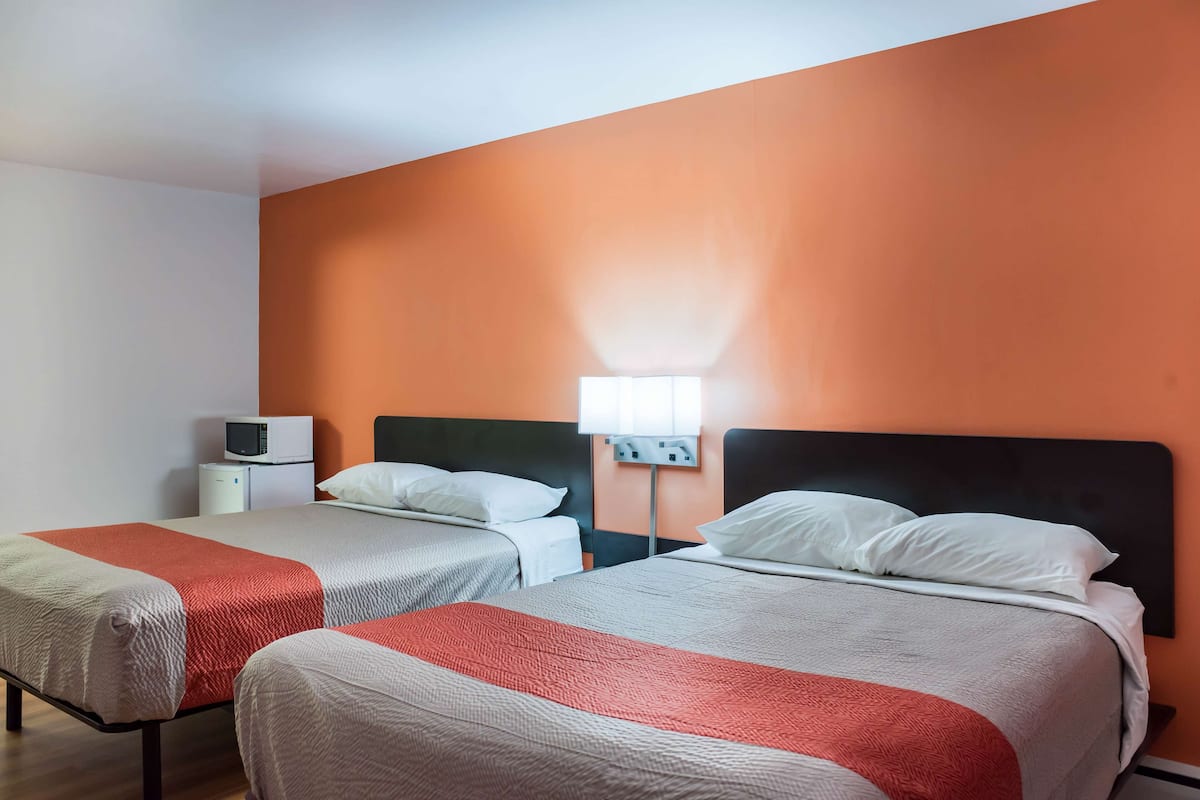 Habitación de hotel con dos camas, cada una con cubrecama rojo y blanco, una mesita de noche blanca, una lámpara blanca y un microondas en la pared.