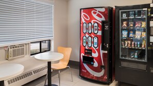 Una macchina distributrice con il logo Coca-Cola, un tavolo e una sedia in una stanza con una finestra e persiane.