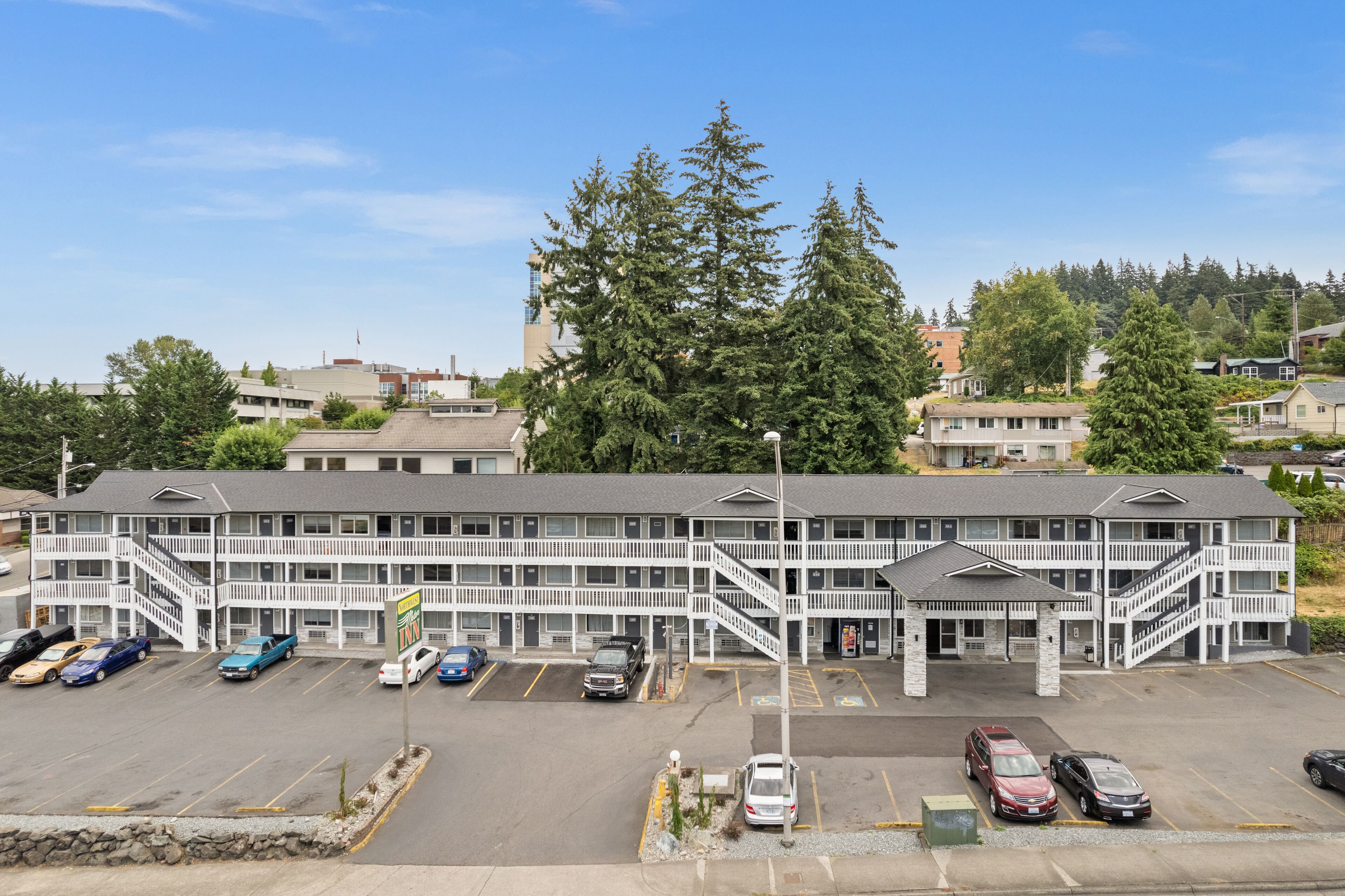 A multi-story motel with a parking lot, outdoor seating area, and a large tree in the background.