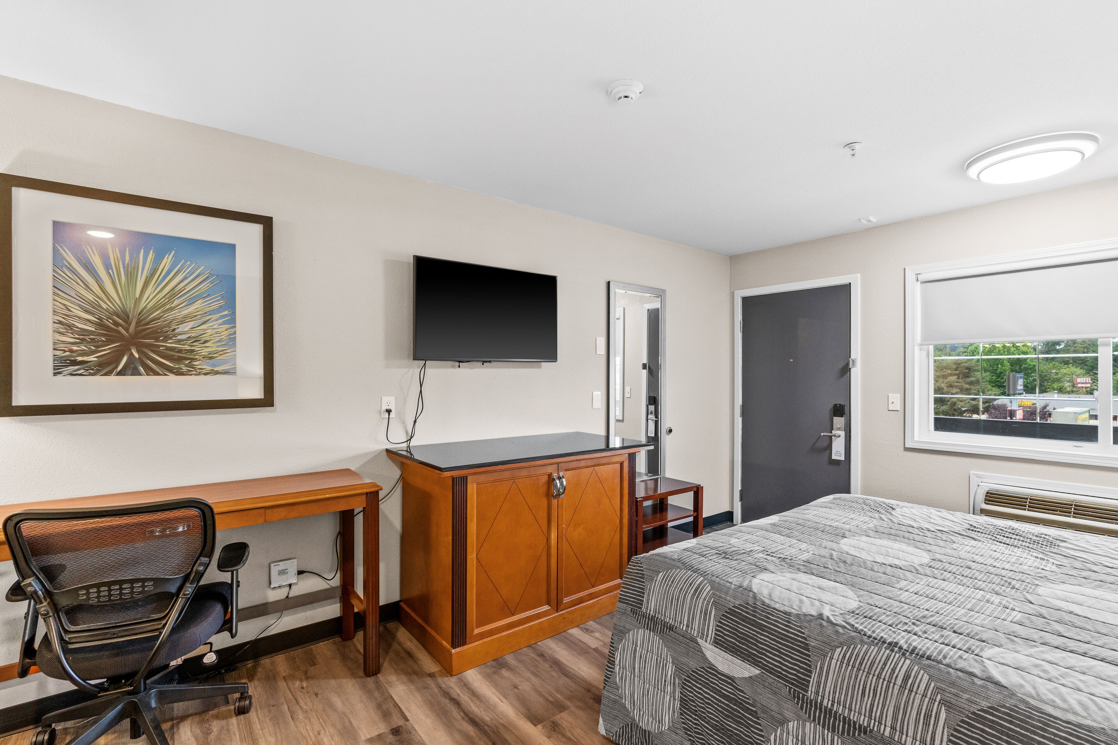 A hotel room with a wooden desk, a bed with a patterned bedspread, a mounted TV, and a framed picture on the wall.