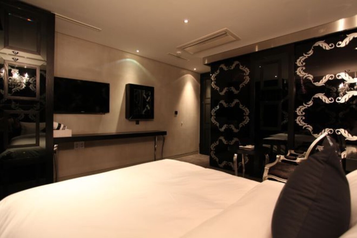 Habitación de hotel con una cama grande, un televisor de pantalla plana montado en la pared, un espejo con diseños ornamentales y una araña.