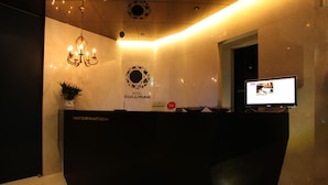 A hotel reception desk with a modern design, a chandelier, a television screen, and a sign indicating the hotel name.