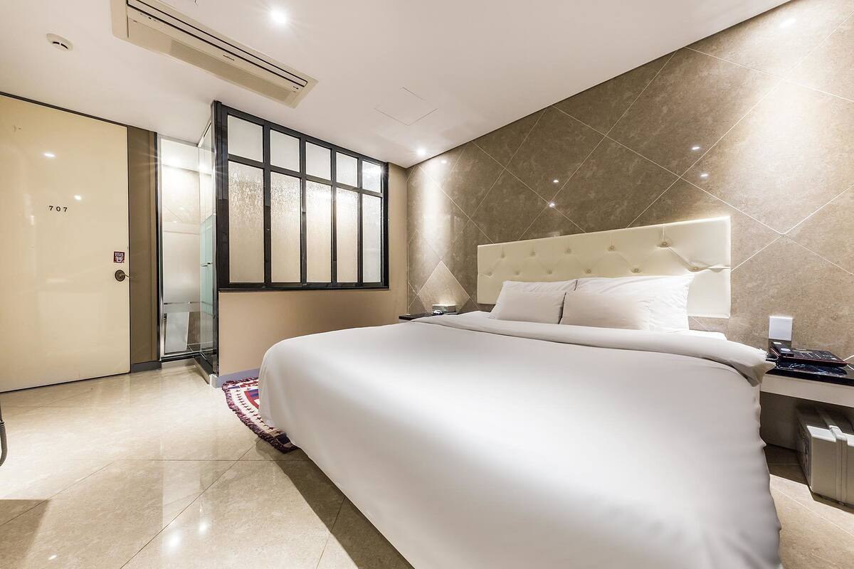 Een moderne hotelkamer met een groot bed, een nachtkastje, een glazen deur naar een badkamer en uitzicht op de stad door een groot raam.