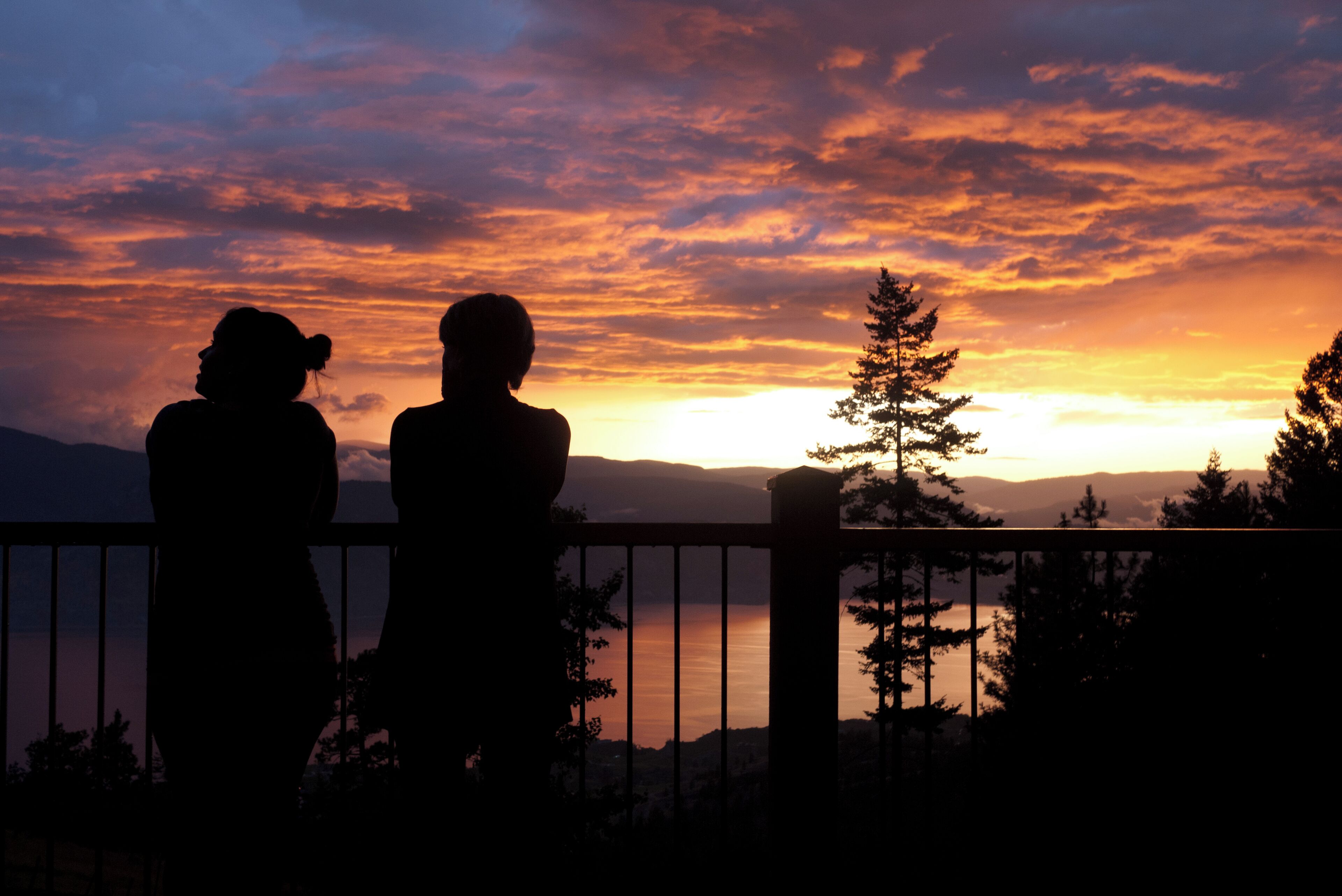 Two silhouettes of people standing on a balcony overlooking a sunset with a lake and mountains in the background.