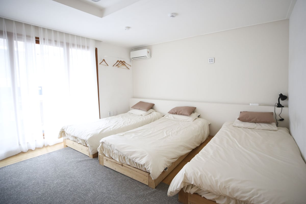 A hotel room with two single beds, a large window with sheer curtains, an air conditioning unit, and a bedside table with a lamp.
