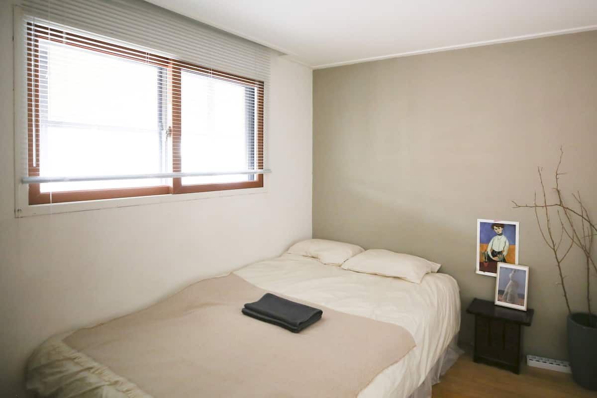 A bedroom with a bed, a window with blinds, a bedside table with a picture frame, and a potted plant.