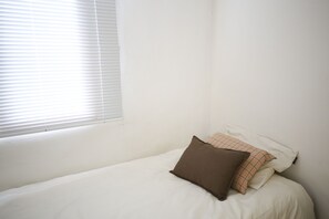 Una cama individual con ropa de cama blanca y dos almohadas, una marrón y otra de cuadros, en una habitación con paredes blancas y una ventana con persianas.
