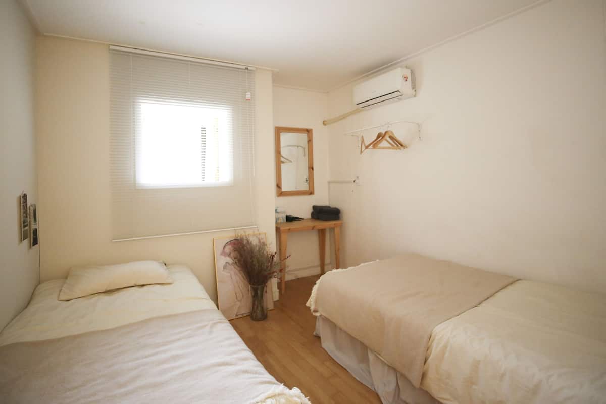 A small, clean room with two beds, a window with blinds, a mirror, a desk, and an air conditioning unit.