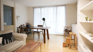 Una sala de estar con una mesa de comedor de madera, un sofá, una mesa de centro y un taburete de madera pequeño. Hay una ventana con cortinas translúcidas y una lámpara sobre un trípode.