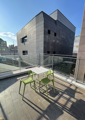A rooftop terrace with a modern building in the background, a glass railing, and a table with chairs.