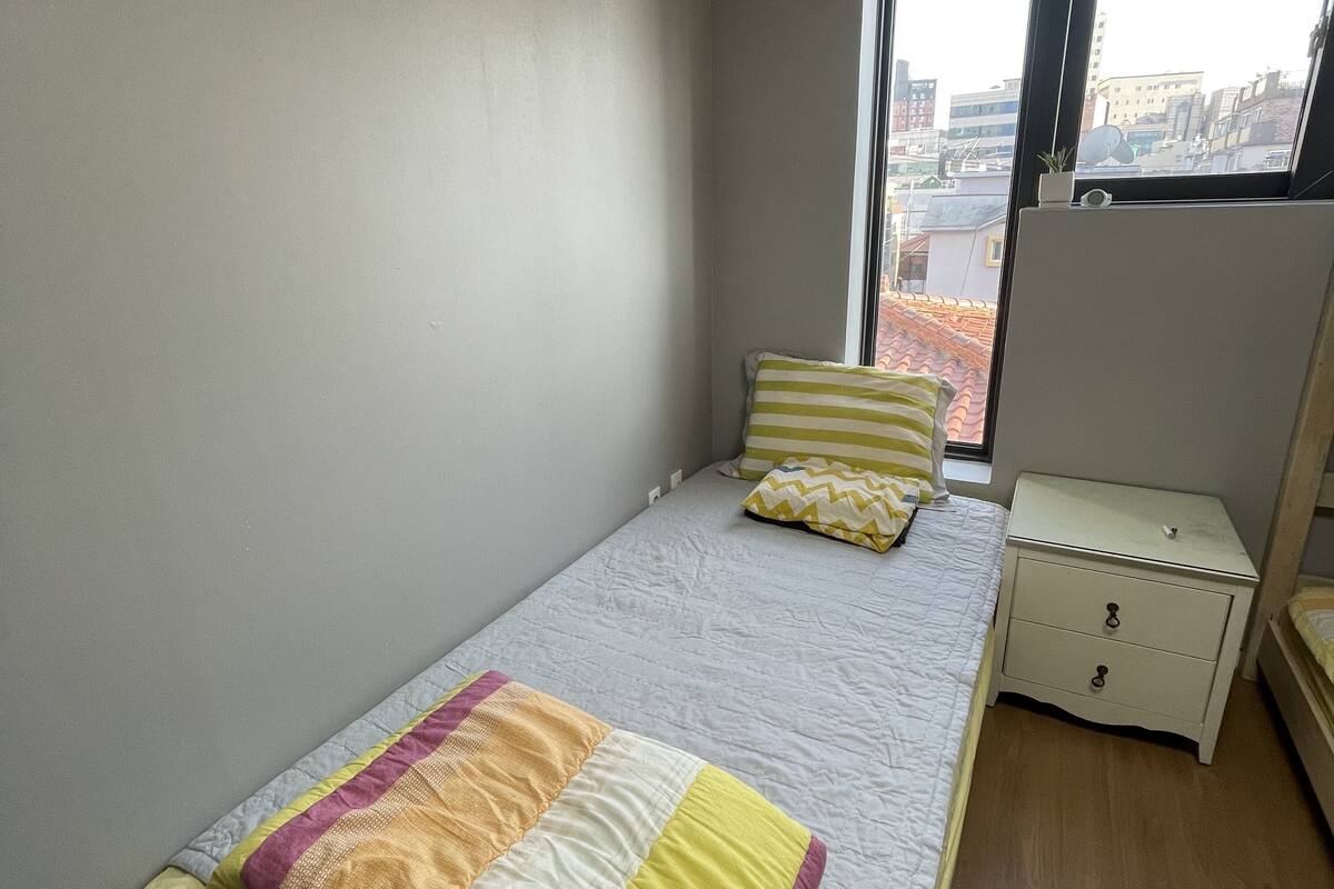 Ein kleines, ordentlich eingerichtetes Schlafzimmer mit einem Einzelbett, einem Nachttisch und einem Fenster mit Blick auf Gebäude.