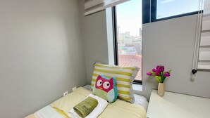 A neatly made bed with a green towel, a pillow with an owl design, and a striped pillow. A vase with purple flowers is on a white dresser next to the bed. - Dream Comfort (Seoul)