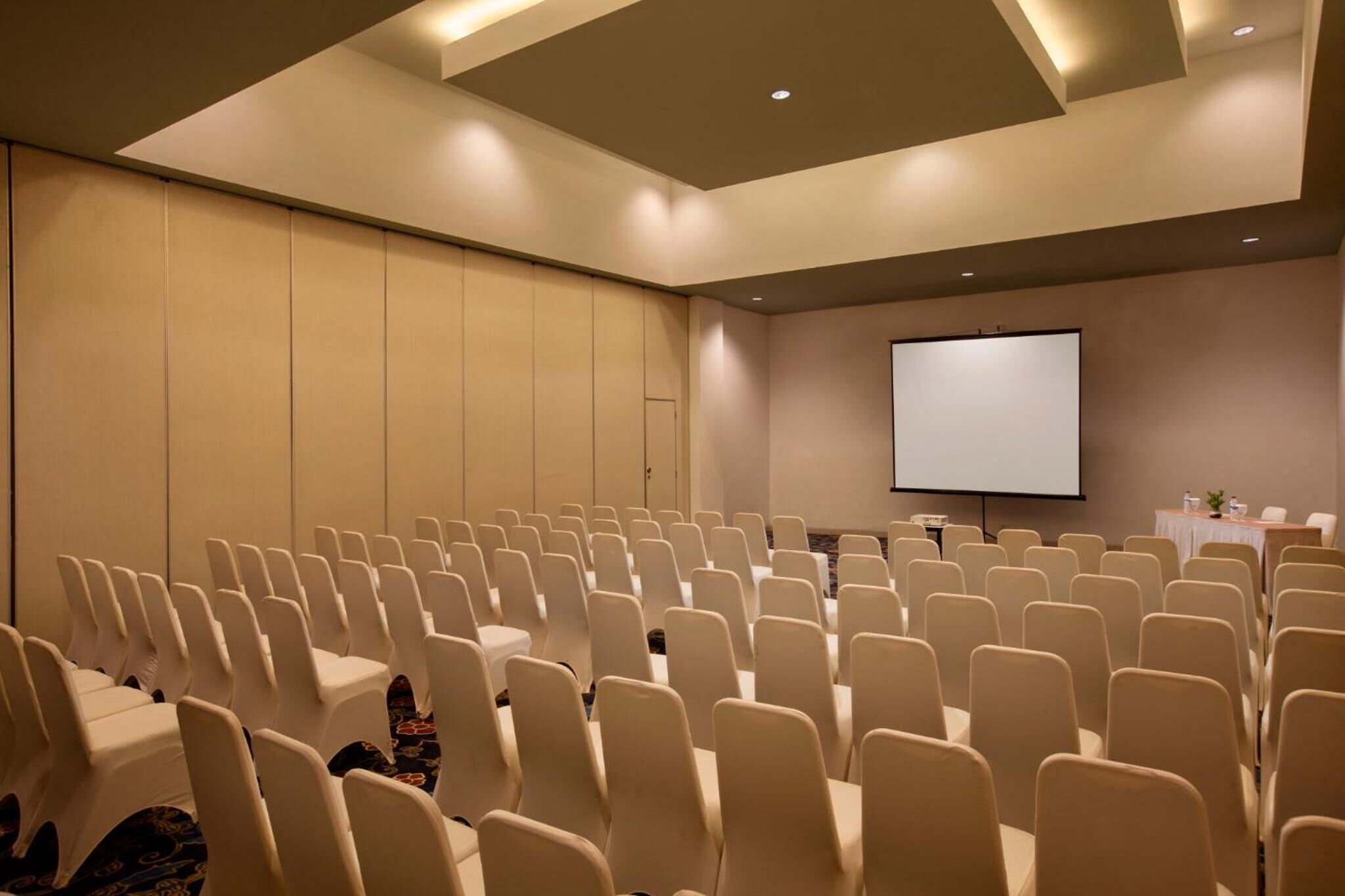 Une salle de conférence avec des rangées de chaises, un écran de projection et une table ornée d’une plante.