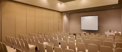 Un salón de conferencias con filas de sillas, una pantalla de proyección y una mesa con una planta.