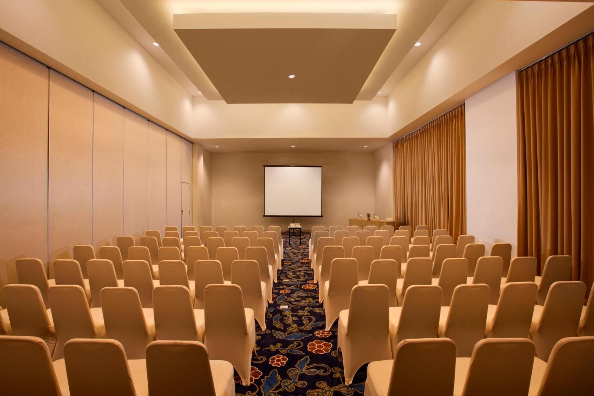 Une salle de conférence avec des rangées de chaises disposées face à un écran de projection, un sol recouvert de moquette et une décoration aux couleurs neutres.