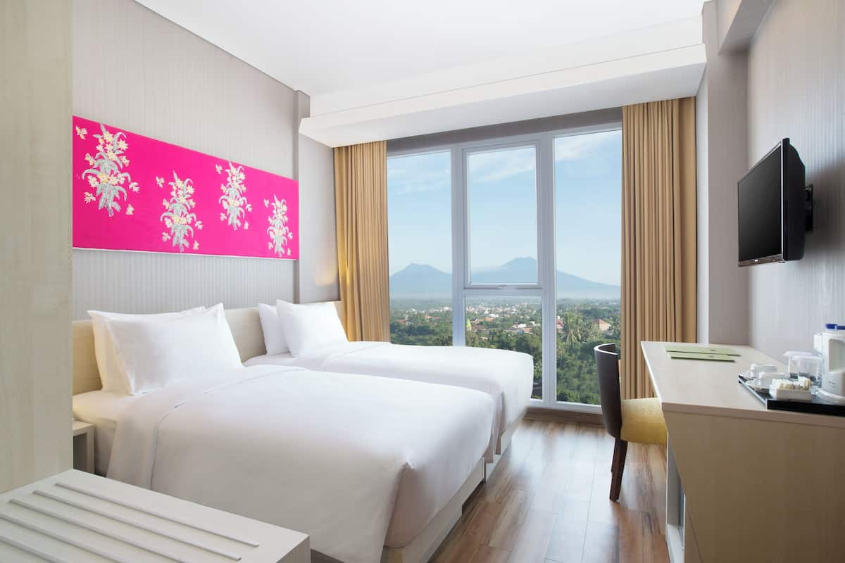 A hotel room with two beds, a desk, a TV, and a large window with a view of the city and mountains.