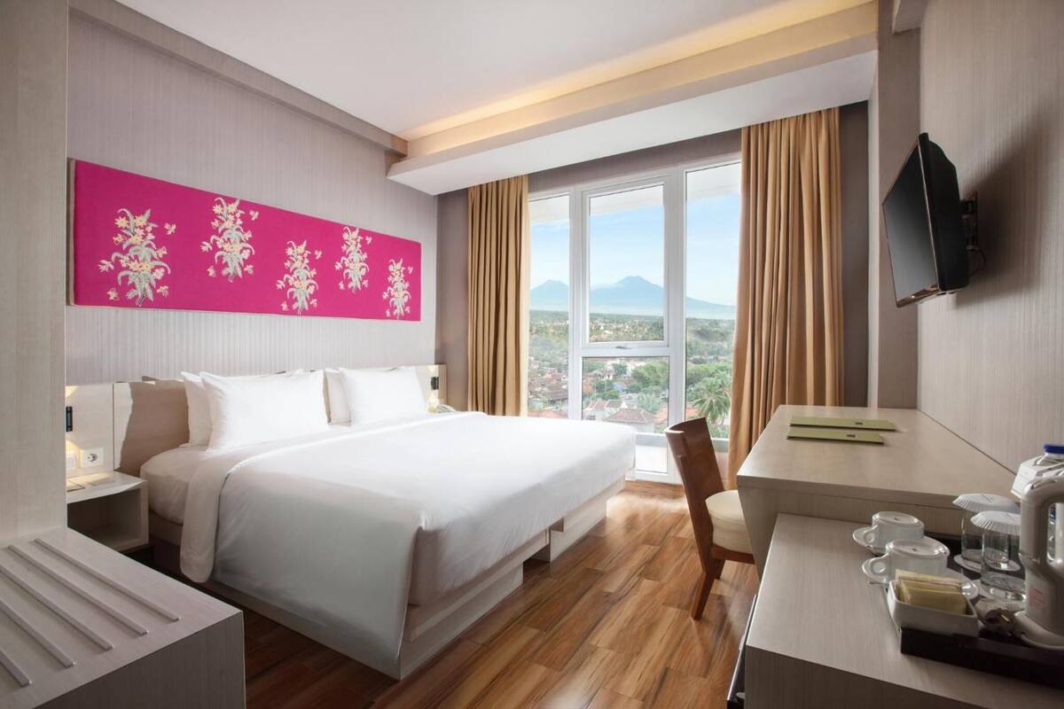 A hotel room with a large bed, a desk with a chair, a television, and a window with a view of a city and mountains.