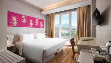 A hotel room with a large bed, a desk with a chair, a television, and a window with a view of a city and mountains.