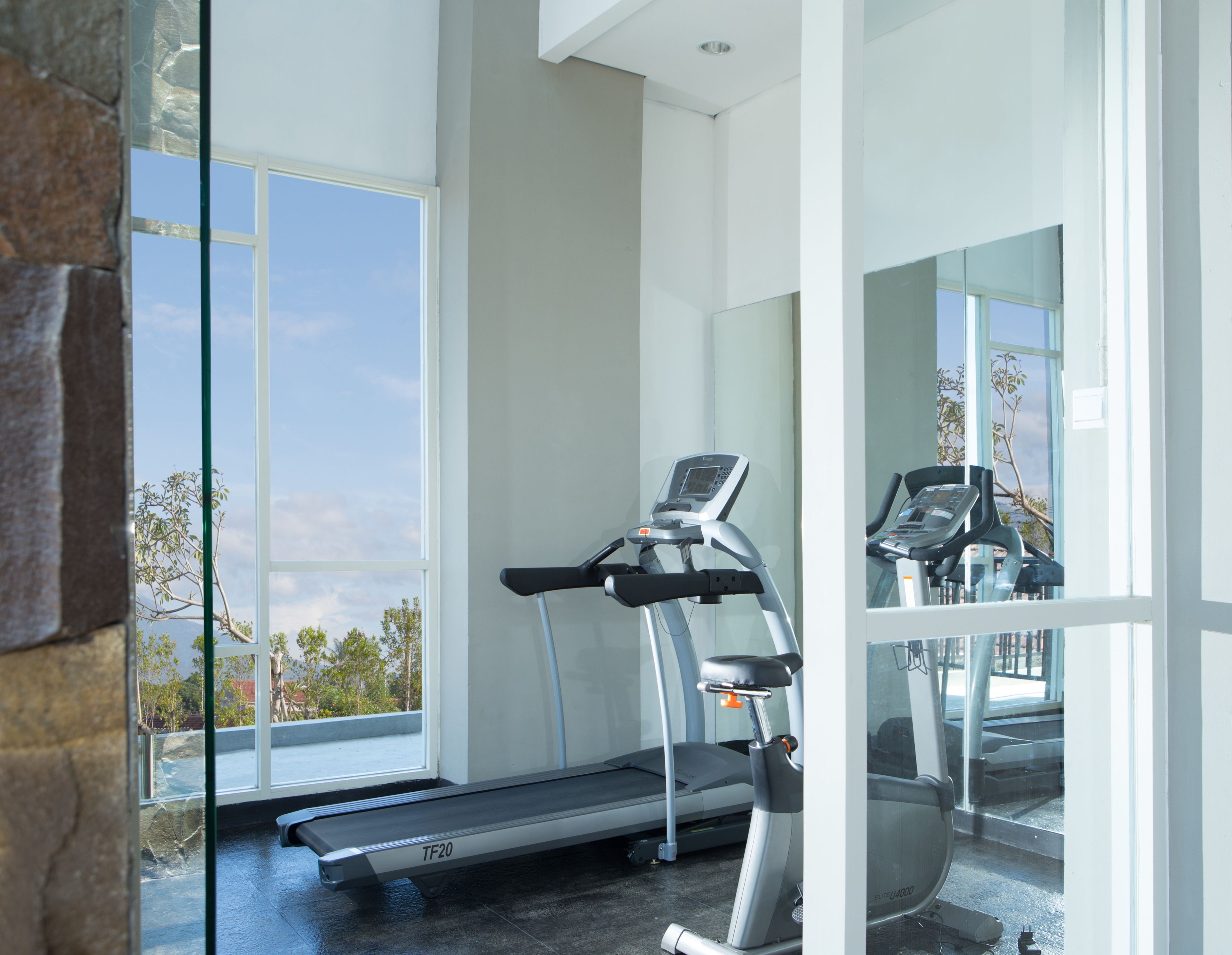 A modern gym with a treadmill and a stationary bike, large windows offering a view of the outdoors.