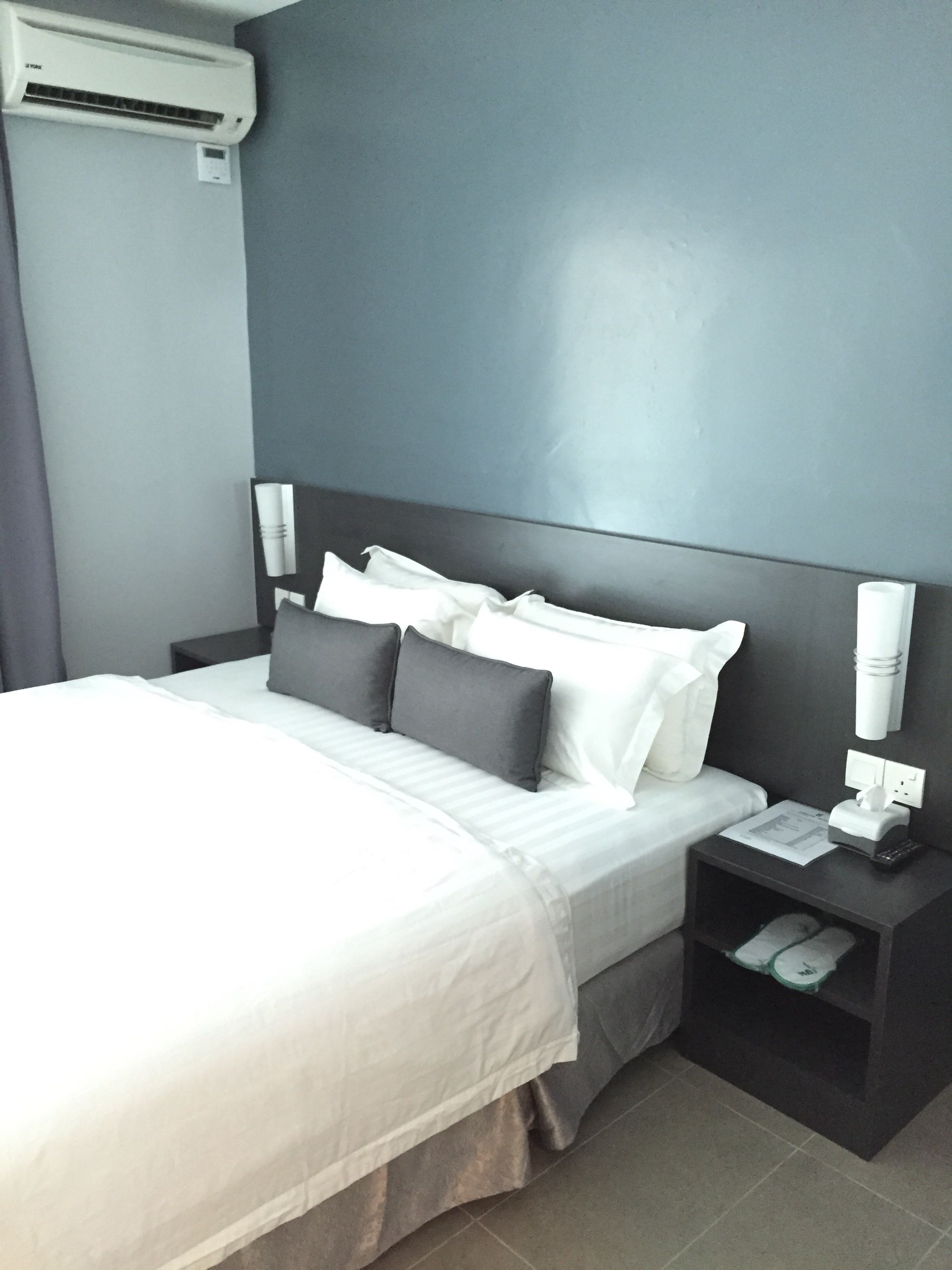 Quarto de hotel moderno com uma cama grande, paredes cinzentas e uma mesa de cabeceira com abajur e telefone.