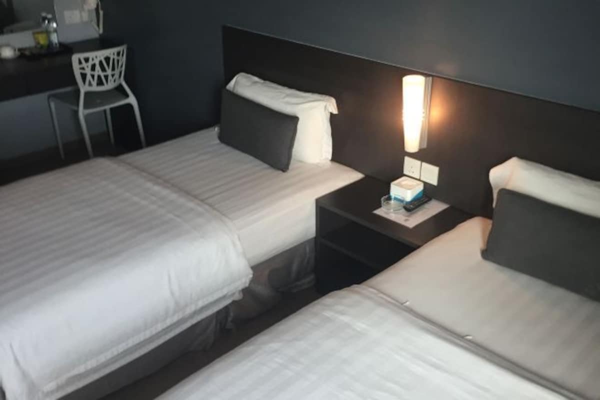 A hotel room with two beds, a bedside table with a lamp, a chair, and a small desk area.