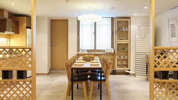 Un comedor con una mesa preparada para cuatro personas, rodeada de sillas de madera. Hay una ventana con persianas, un armario con vajilla y una lámpara de techo decorativa.