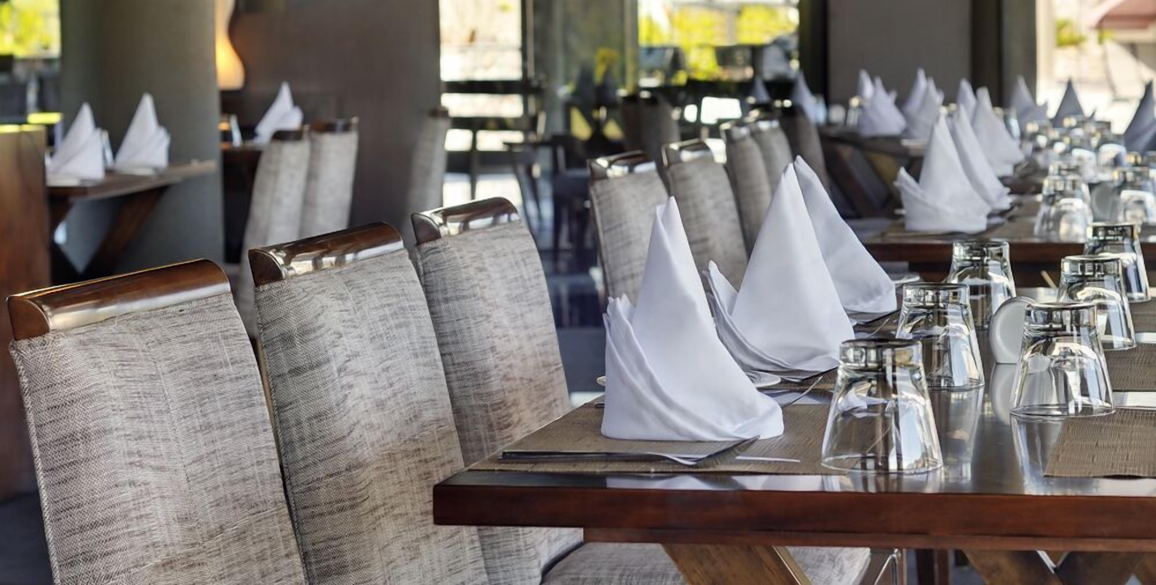 A restaurant with neatly arranged tables, each with a folded white napkin, clear glassware, and a dark wooden table surface.