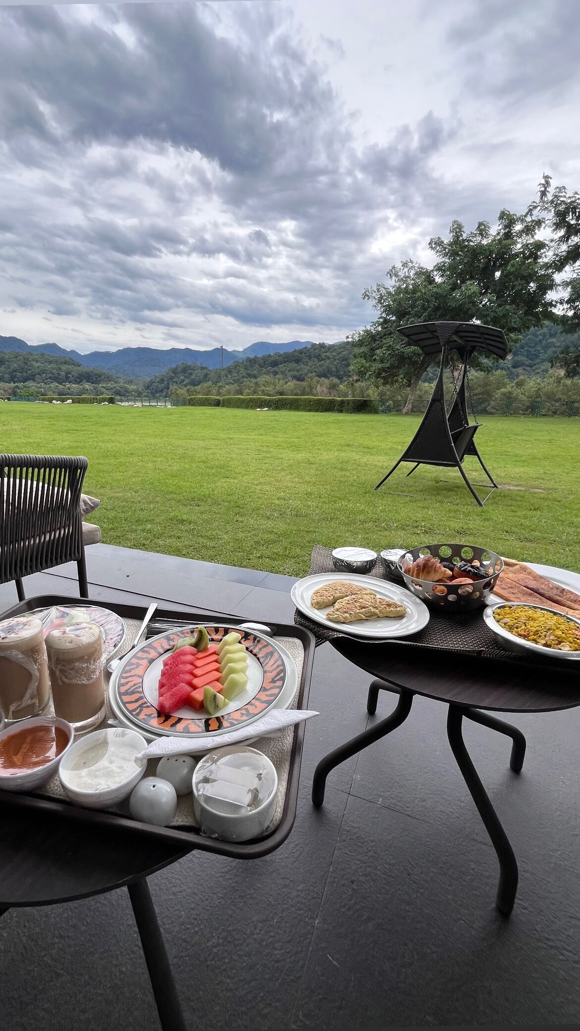 Mesa al aire libre dispuesta para dos personas, con una variedad de platillos, incluyendo frutas, pastelería y una parrilla.