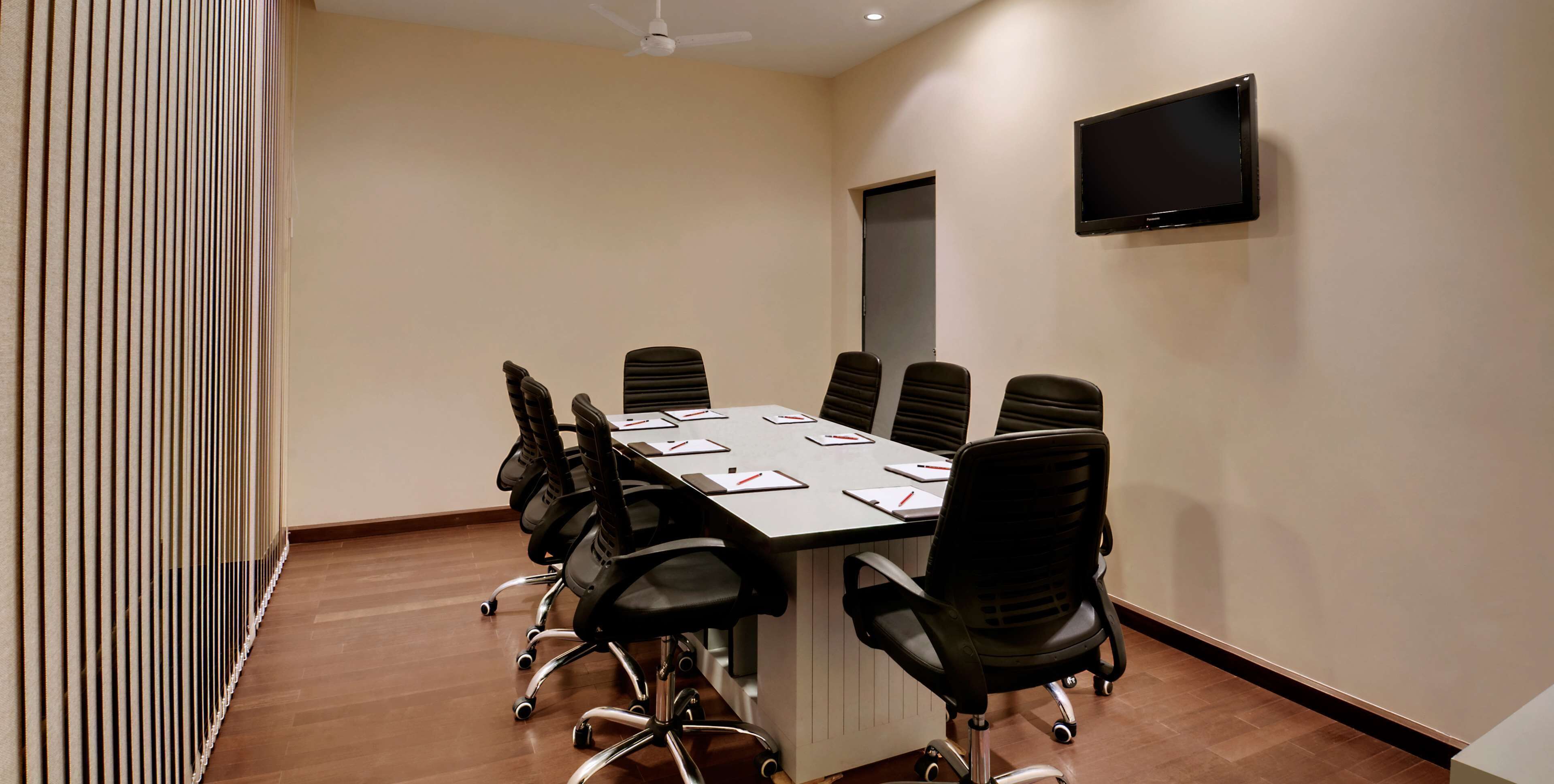 A conference room with a long table, black chairs, a flat-screen TV, and a door.