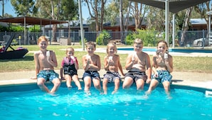 A group of children sitting by a swimming pool with a covered area and trees in the background.