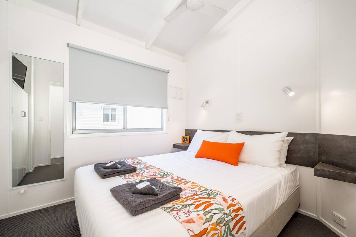 A hotel room with a large bed, a window with a blind, a mirror, a nightstand with a lamp, and a headboard with a built-in shelf.