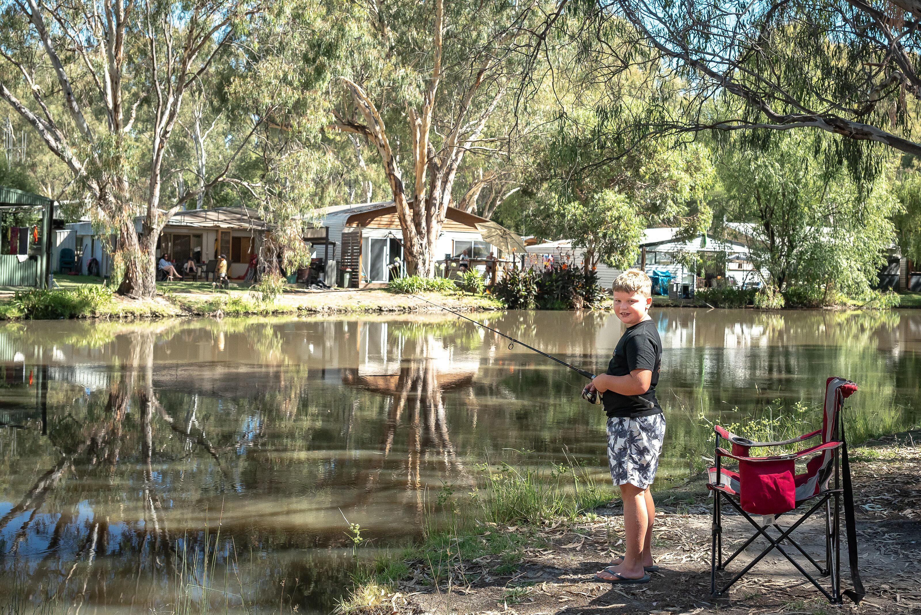 A person fishing by a calm lake with trees and houses in the background.