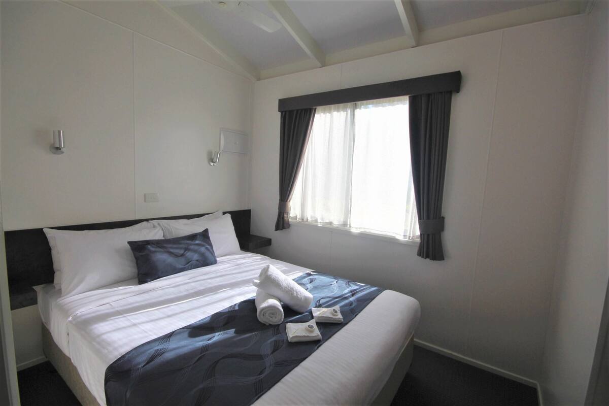 A neatly made bed with white linens, a dark blue blanket, and two rolled towels.