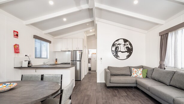 A modern living room with a grey sofa, a dining table with chairs, and a kitchen in the background.