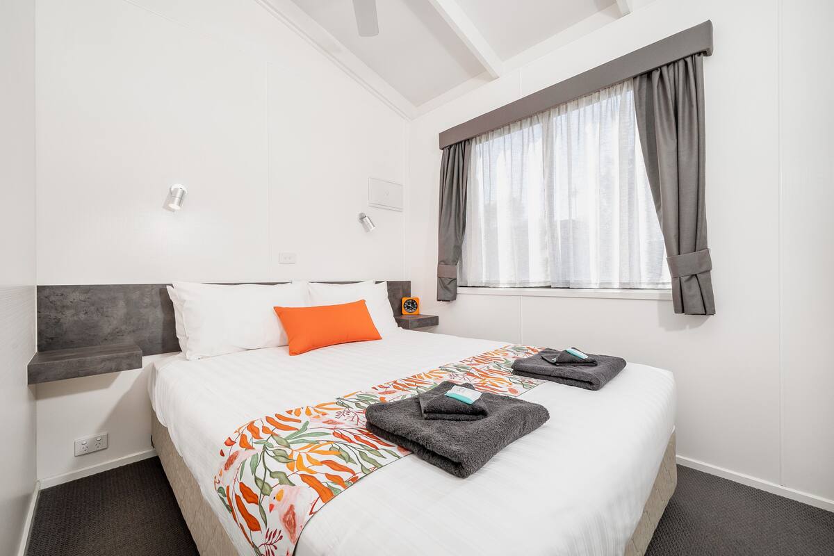 A hotel room with a large bed, a bedside table, a nightstand, a window with curtains, and a ceiling fan.