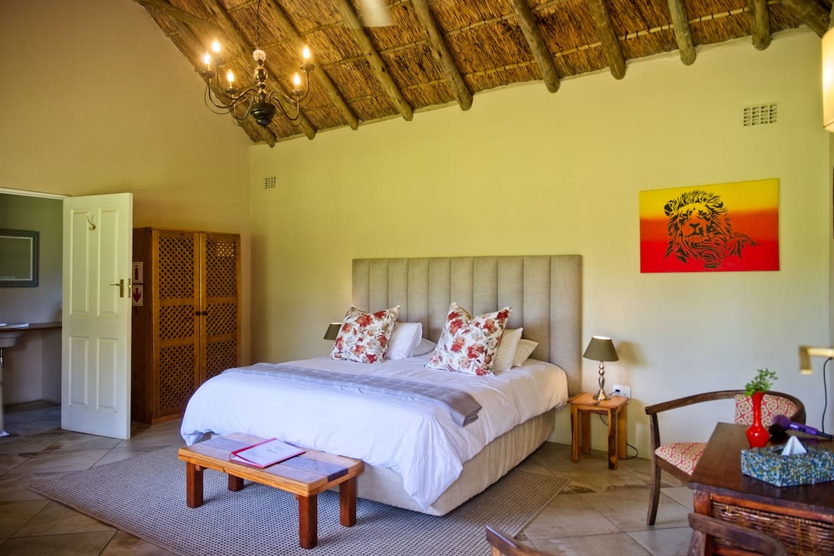 A bedroom with a large bed, a wooden headboard, a bedside table with a lamp, a chair, and a painting on the wall.