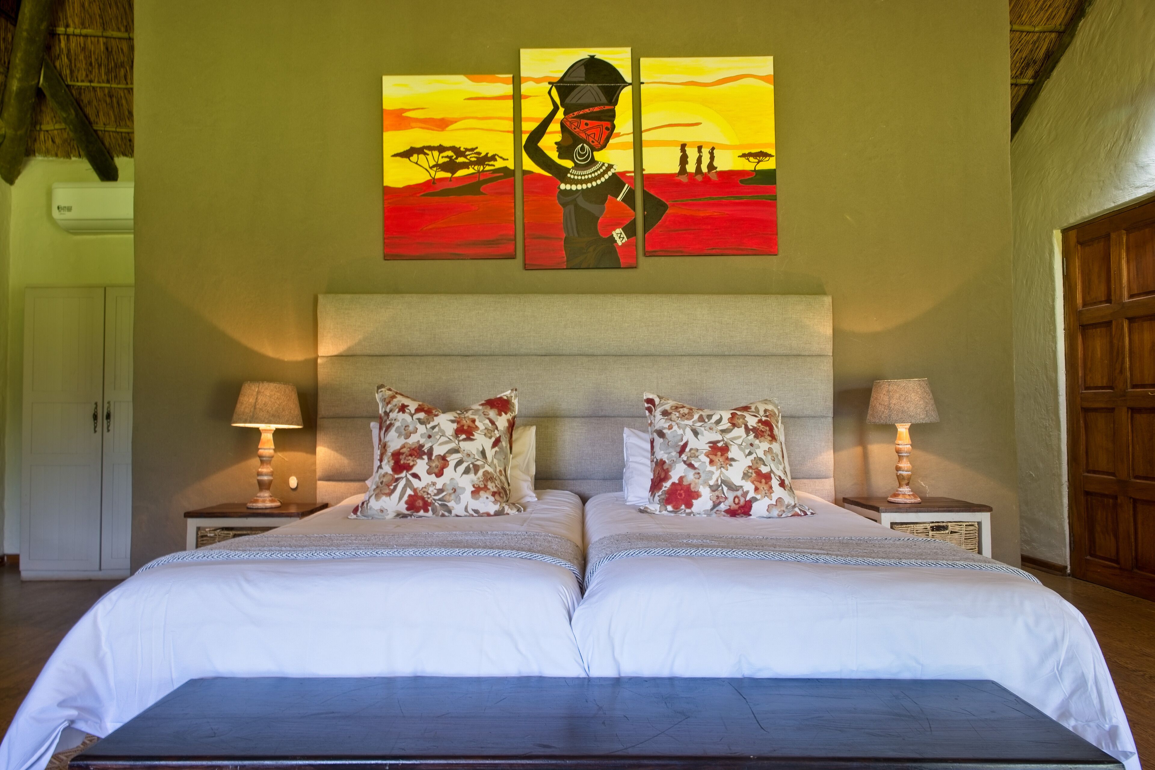 Una cama doble con ropa blanca de cama y almohadas florales, una mesita de noche con lámpara y un cuadro en la pared de una figura con traje tradicional.