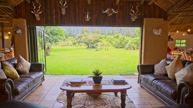 A spacious room with a large window offering a view of a well-maintained lawn. There are two leather sofas with cushions, a wooden coffee table, and a rug on the floor. - Chrislin African Lodge (Addo)