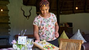 A person serving food on a wooden board at a table with a number 3 sign, in a room with a rustic interior and a leather couch. - Chrislin African Lodge (Addo)