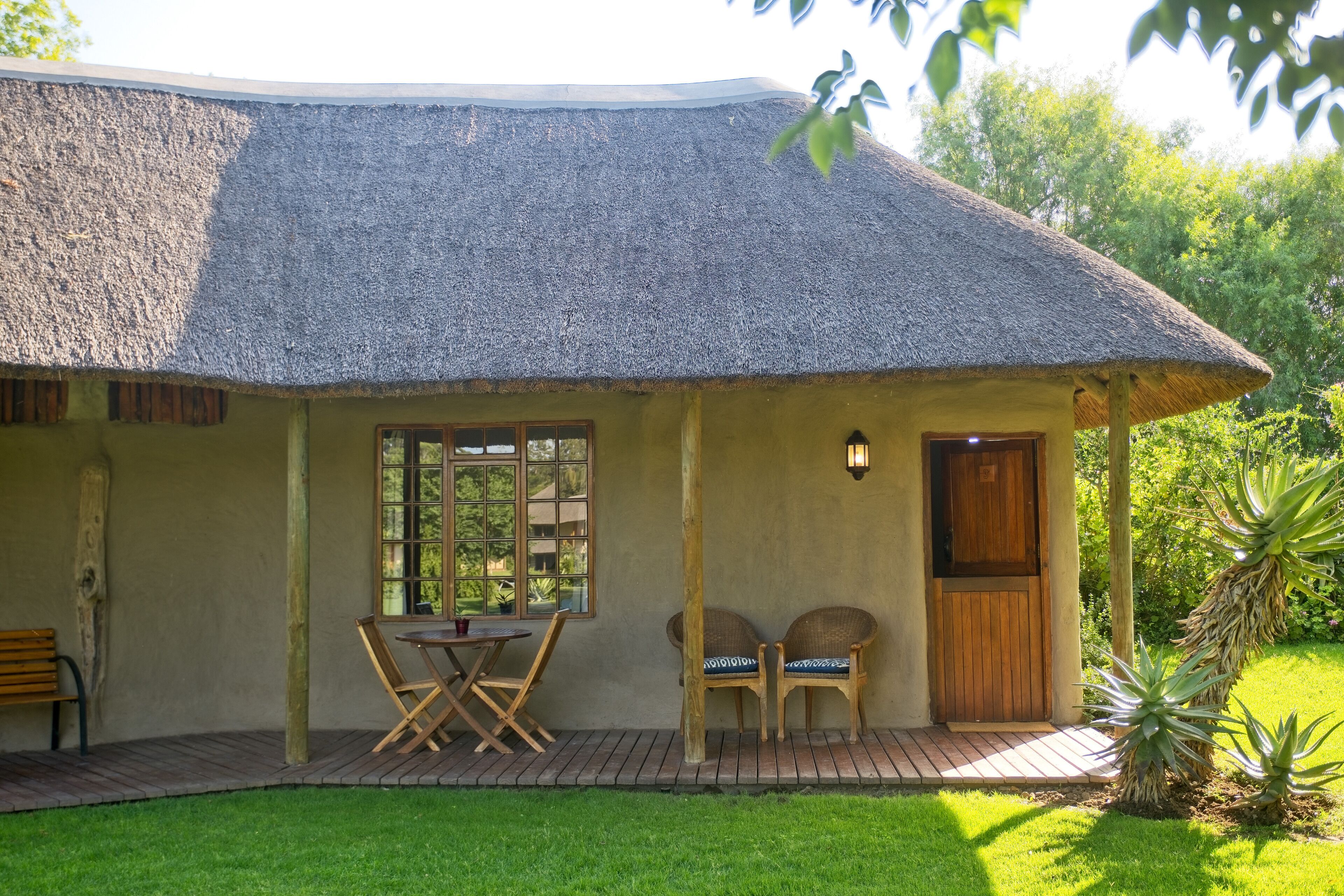 A thatched-roof house with a wooden deck, two wicker chairs, a table, and a bench.