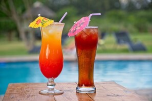 Deux boissons, chacune agrémentée d’un petit parapluie, sont posées sur une table en bois, près d’une piscine.
