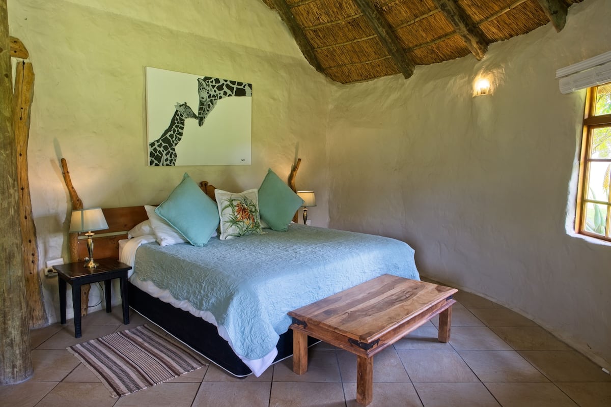 A bedroom with a bed, pillows, a nightstand, a lamp, a wooden bench, a wall art of giraffes, a thatched roof, and a window with a view.