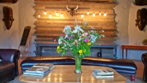 A cozy living room with a wooden ceiling, a fireplace, a sofa, a coffee table with books, and a vase of flowers. - Chrislin African Lodge (Addo)